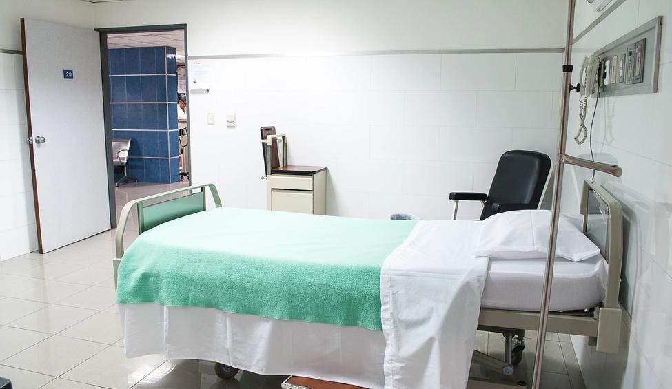 Sykehusseng og medisinsk utstyr med en stol ved siden av. Døren til rommet med sengen er åpent. 