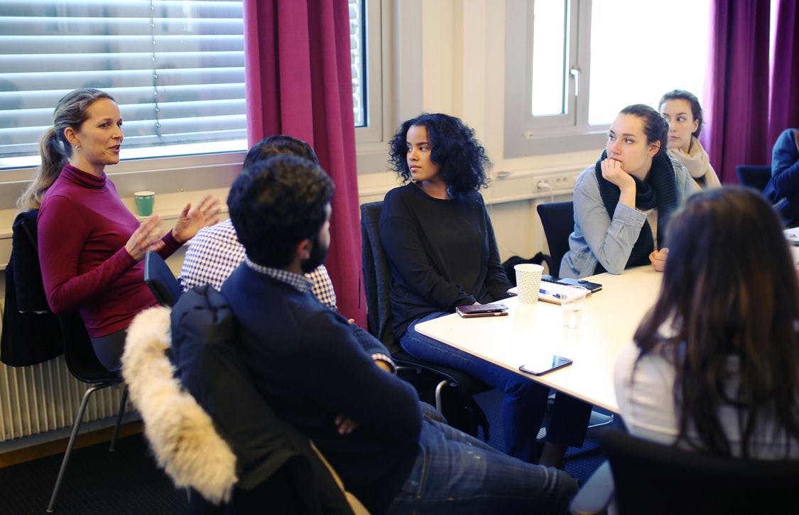 En kvinne foreleser for en gruppe unge mennesker som sitter rundt et bord.