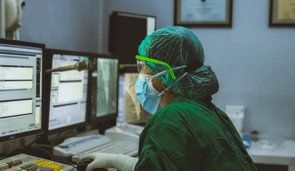 Sykepleier i smittevernutstyr ser mot PC-skjermen
